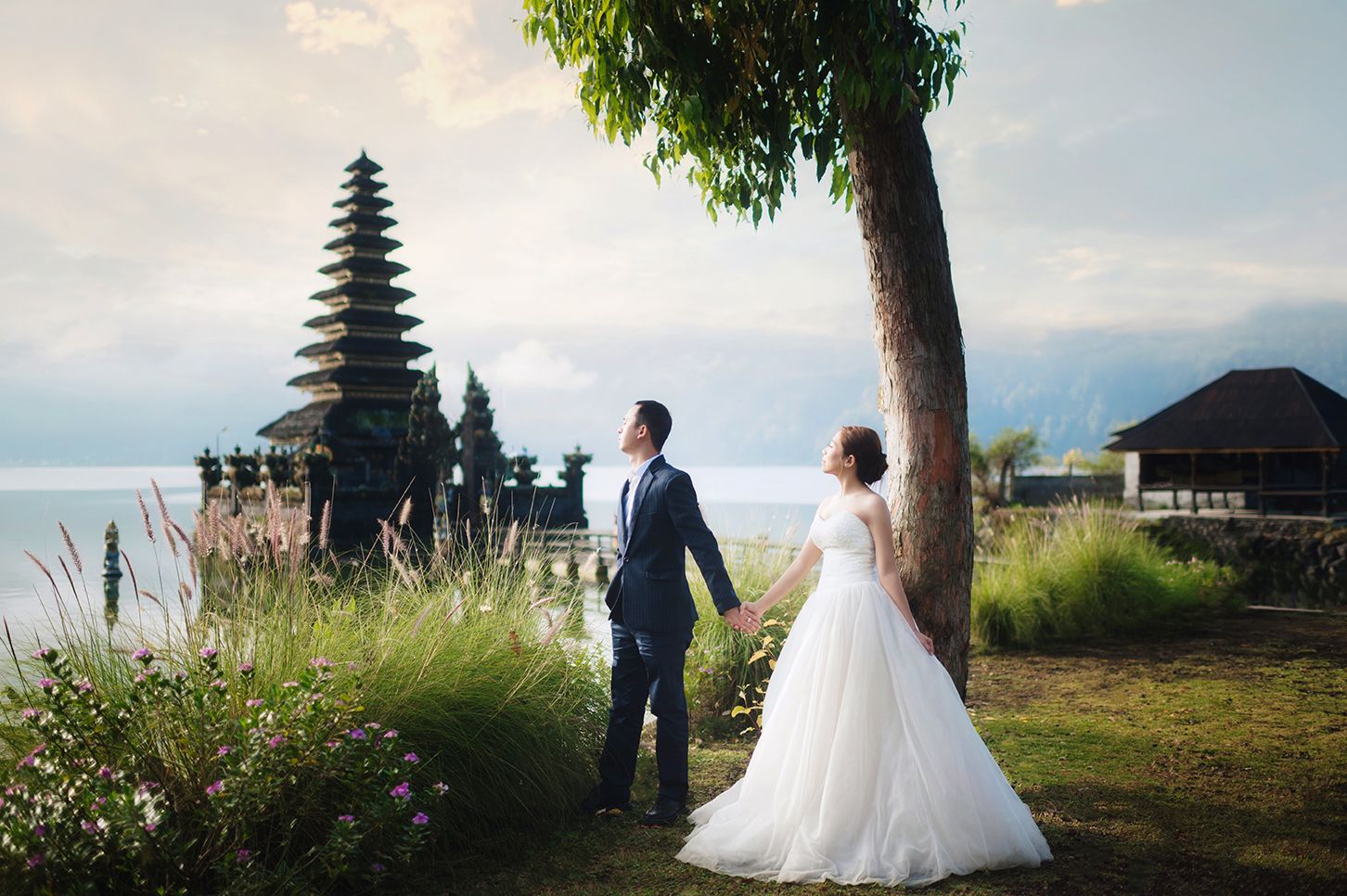 Bride and groom hold hands near a tree with a Balinese temple and lake backdrop on their wedding day.