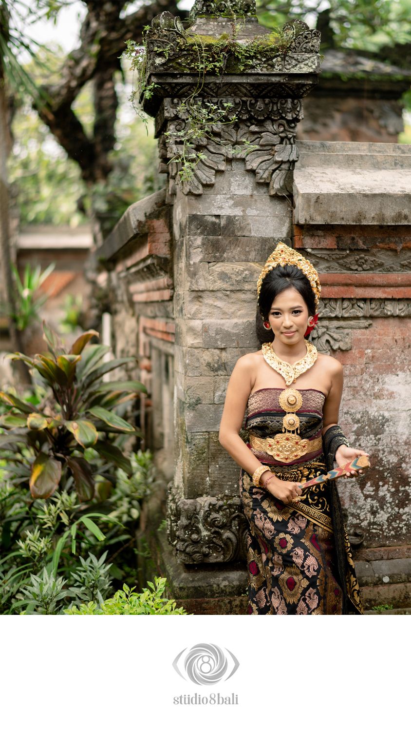 Balinese woman in traditional gold ceremonial dress and headdress poses gracefully before ancient stone temple gates.