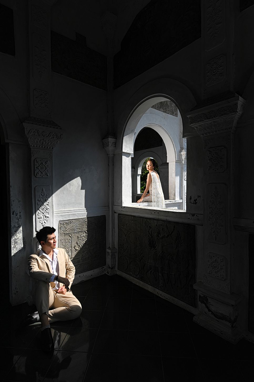 A man in a beige suit sits on the floor while a woman in white stands in an ornate arched window above him.