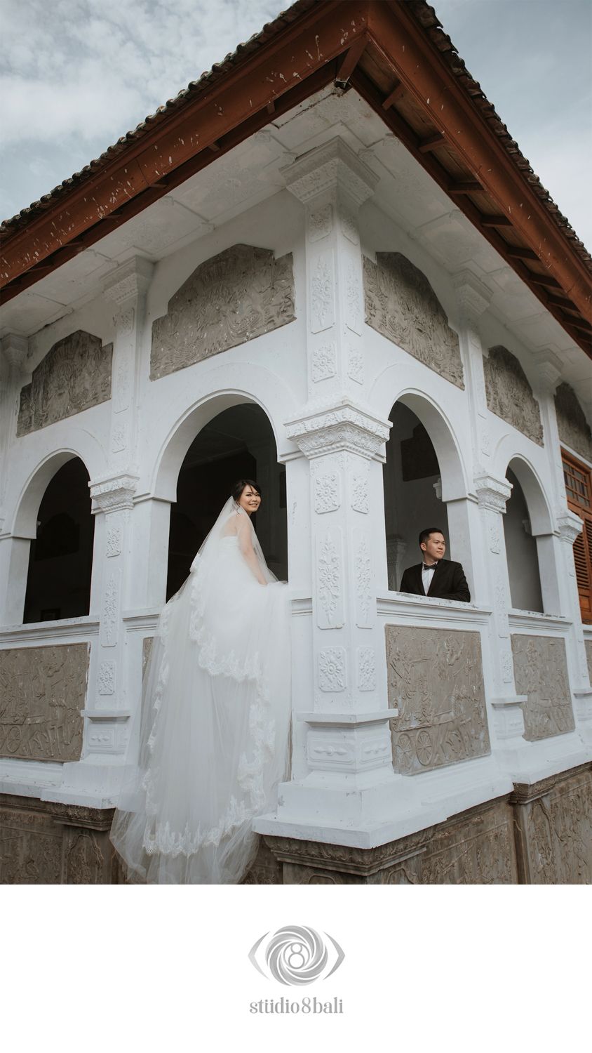 Bride in white gown and groom in tuxedo pose at ornate white arched building for wedding photos.