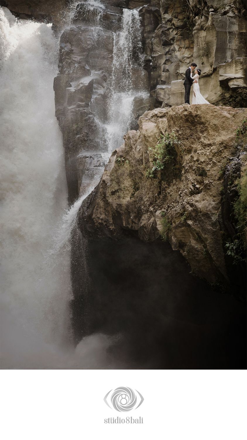Bride and groom standing on rocky cliff beside a dramatic waterfall during their Bali wedding elopement photo session.