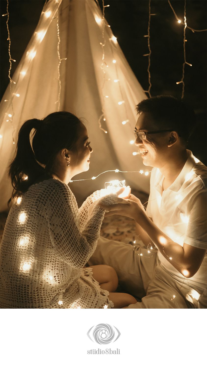 Couple sharing a romantic moment under fairy lights in a cozy tent, holding a glowing jar together.