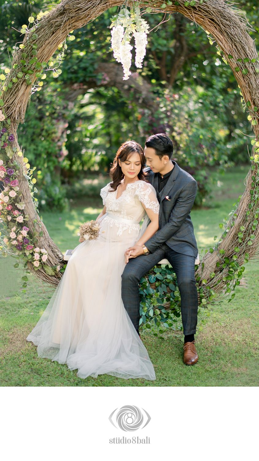 Couple in wedding attire sitting in a floral circular arch surrounded by lush greenery for an elegant outdoor ceremony.