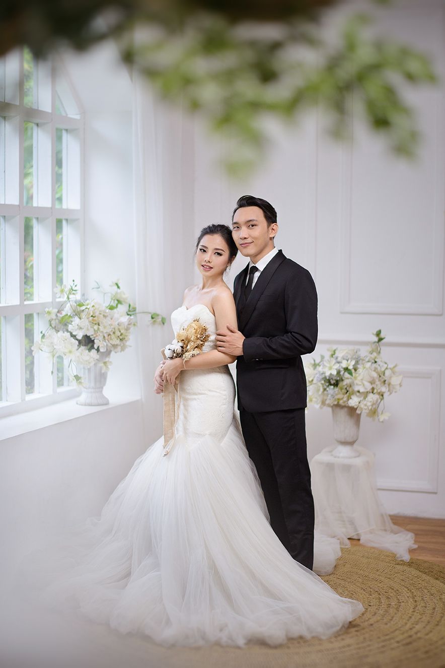 A couple poses in wedding attire, bride in white mermaid gown holding bouquet, groom in black suit, in elegant white studio.
