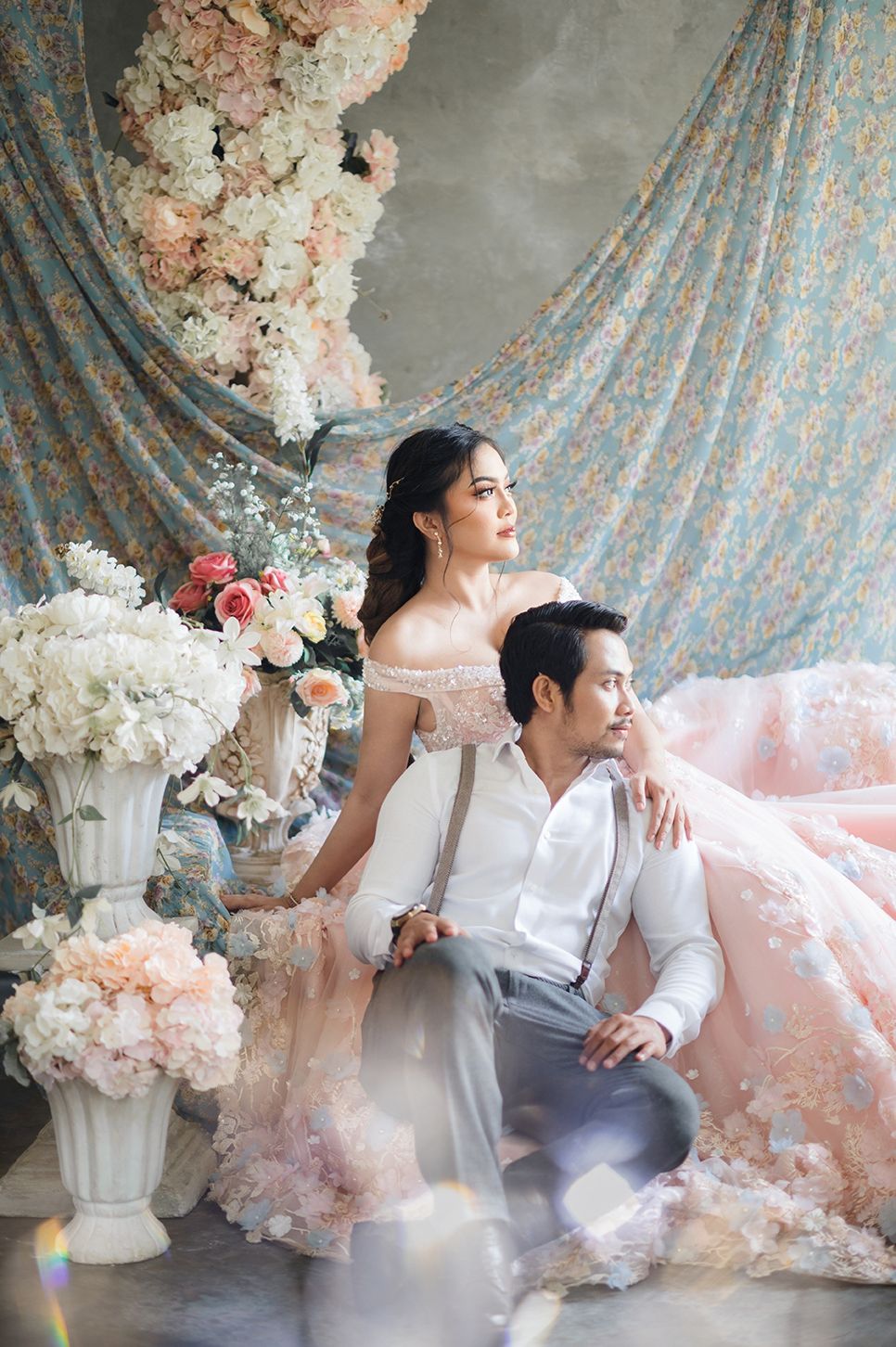 Elegant couple in romantic floral studio setting, woman in pink gown, man in white shirt with suspenders.