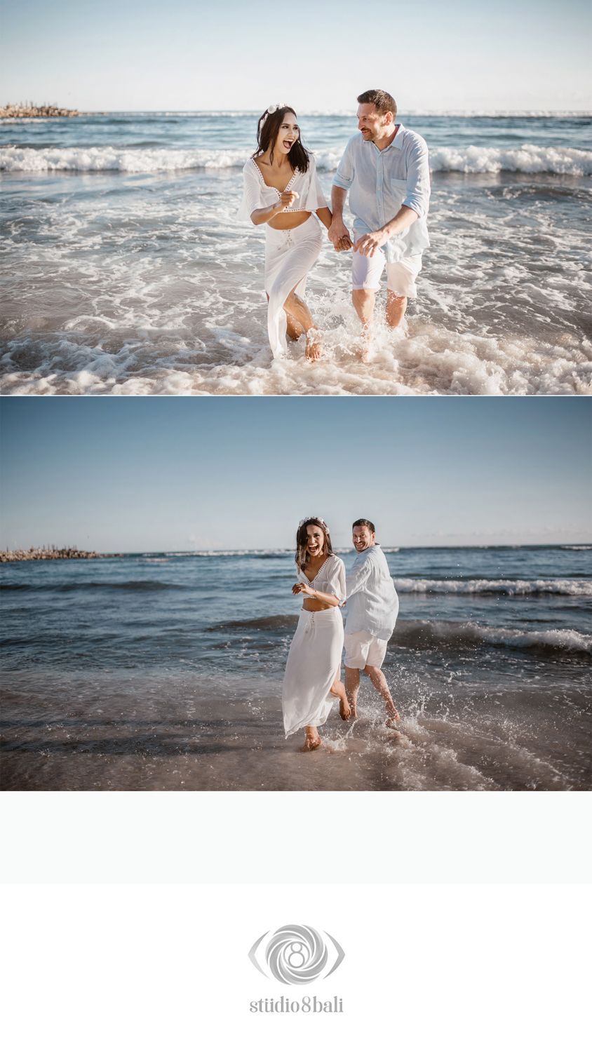 Couple laughing and splashing in ocean waves during beach photo session, both wearing white outfits.