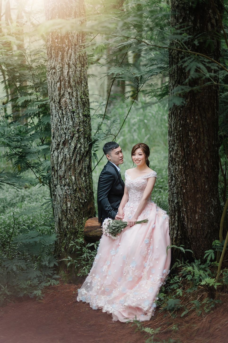 Couple in wedding attire poses romantically among tall trees in a lush forest, bride wearing pink lace gown holding bouquet.