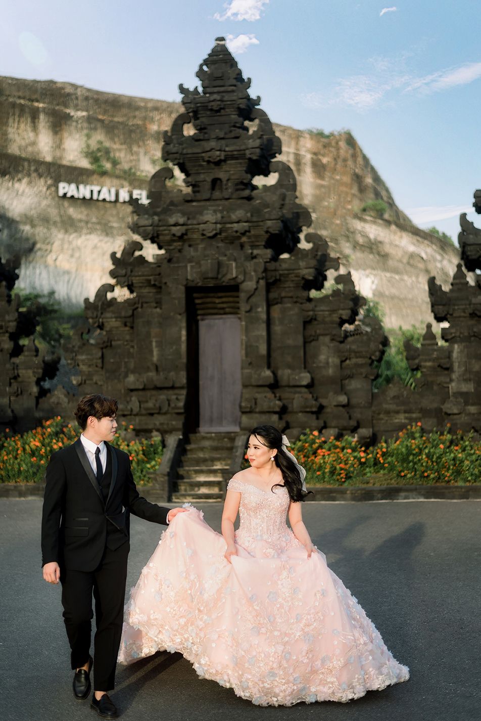 Couple in wedding attire poses before ornate Balinese stone gate, bride in floral gown, groom in black suit.