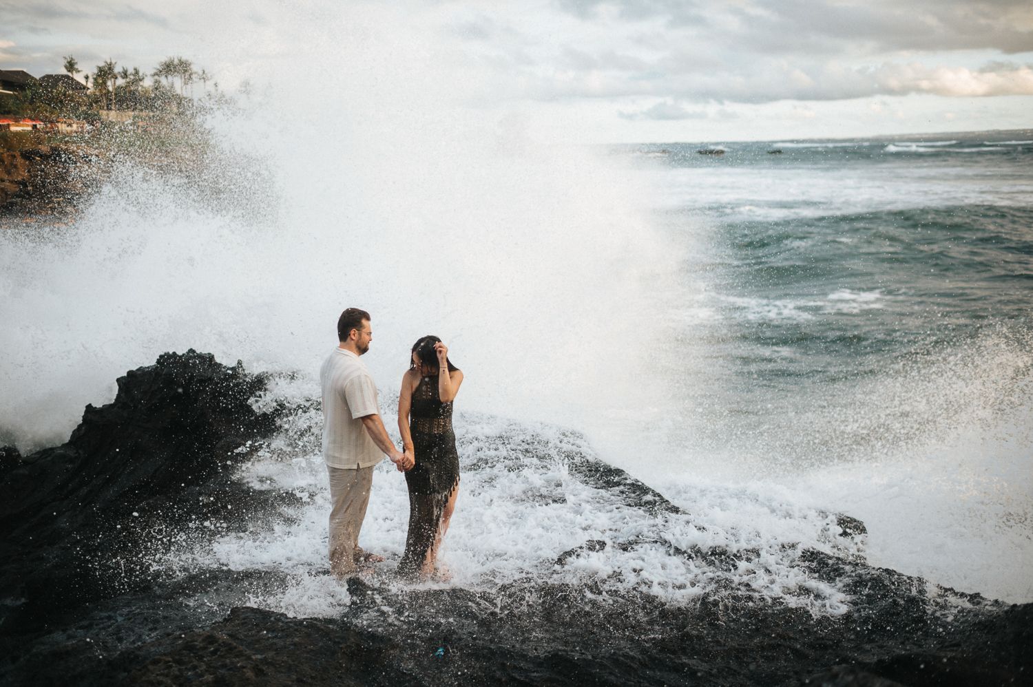 Couple holding hands on rocky Hawaiian shore with dramatic ocean waves crashing behind them.