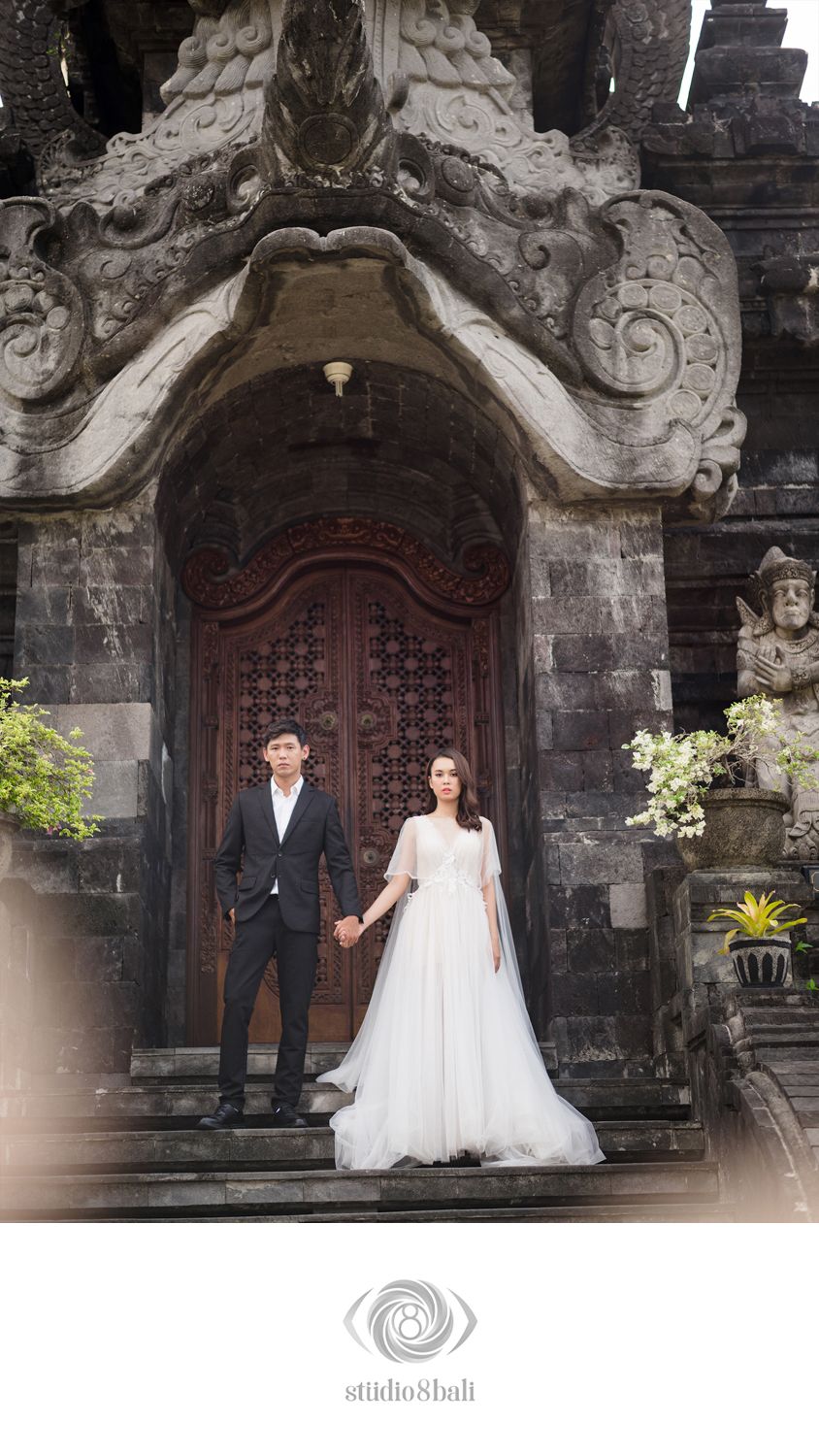 Couple in wedding attire posing before ornate Balinese temple doors, bride in white gown, groom in black suit.
