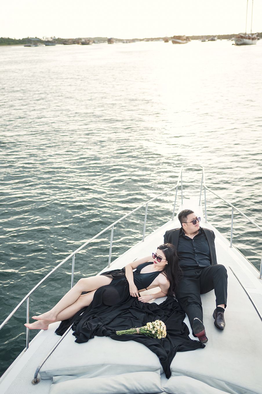 Elegant couple in black attire relaxing on a luxury yacht bow, woman holding white flowers, surrounded by calm ocean waters.