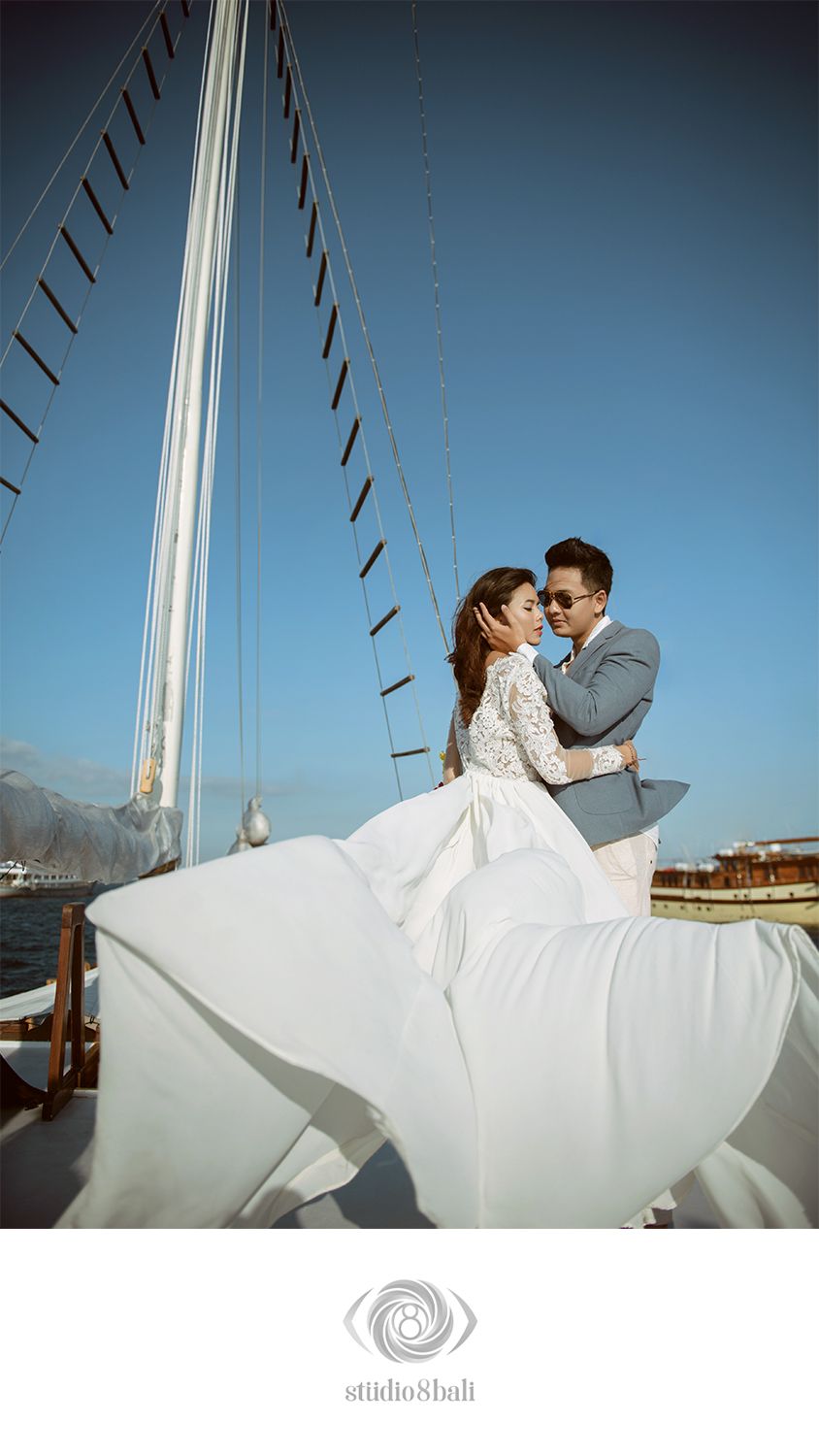 Couple in wedding attire embracing on a sailboat deck under a clear blue sky, bride's white gown flowing in the wind.