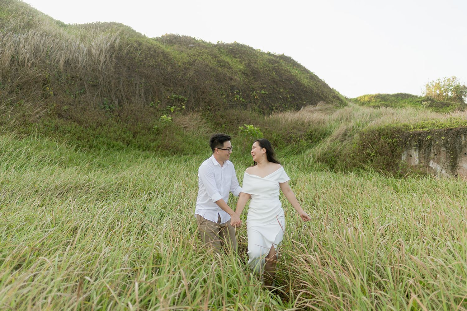 Couple in white outfits holding hands in lush green grass field with scenic hillside backdrop.