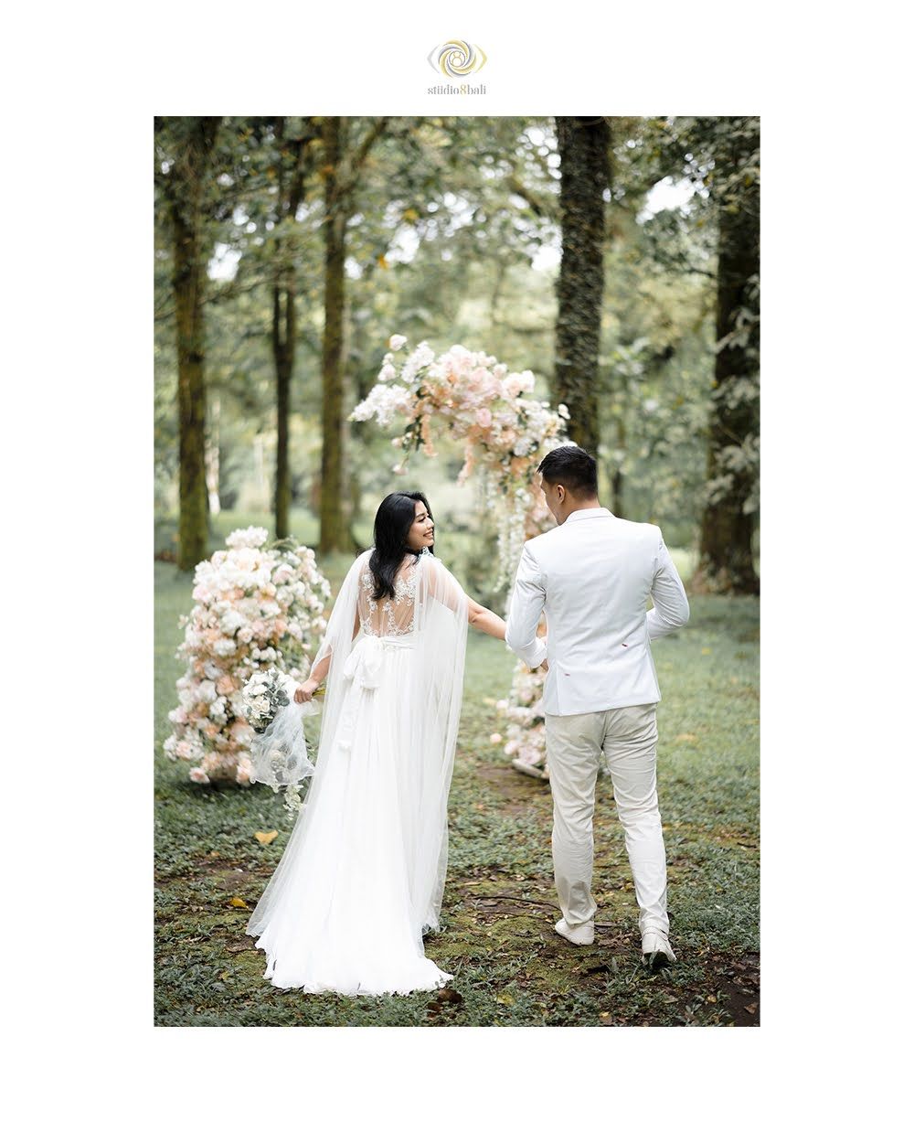 Bride and groom stand before a floral arch in a lush forest setting during an outdoor wedding ceremony.