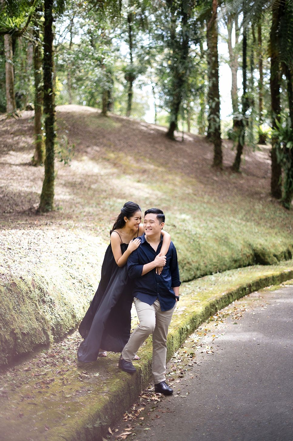 A couple laughing and embracing on a tree-lined path in a lush green park setting.