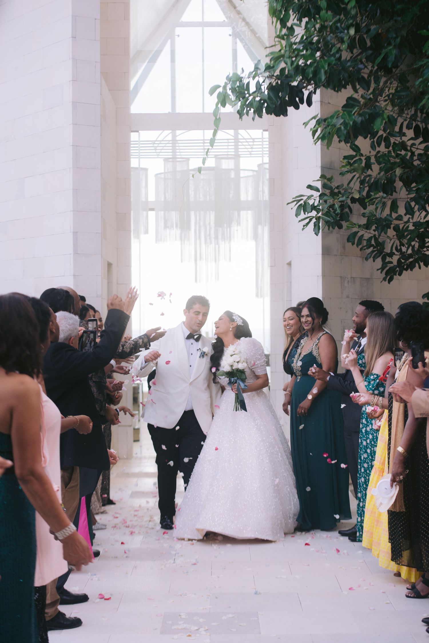 Newlywed couple kissing during wedding recessional as guests toss flower petals in elegant white venue.