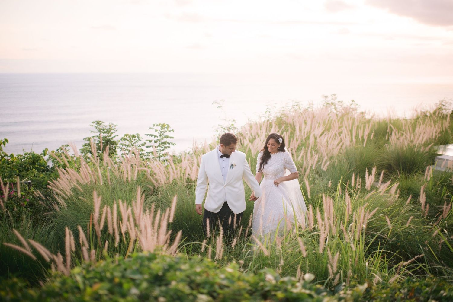 Bride and groom walking through lush green meadow with ocean sunset backdrop on their wedding day.