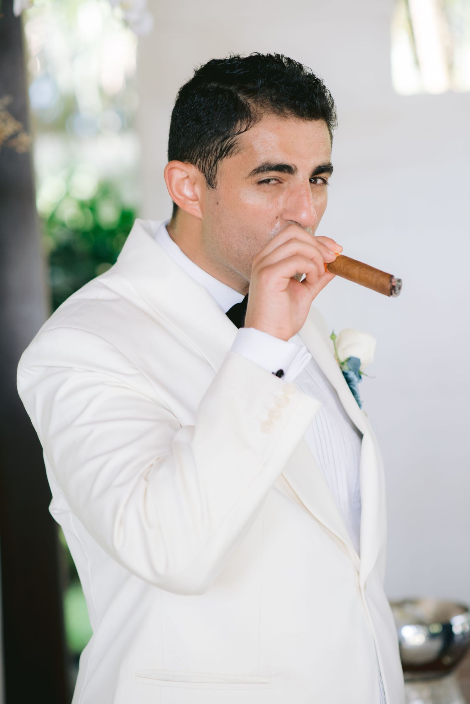 Groom in white tuxedo smoking a cigar on his wedding day, wearing a boutonniere and bow tie.