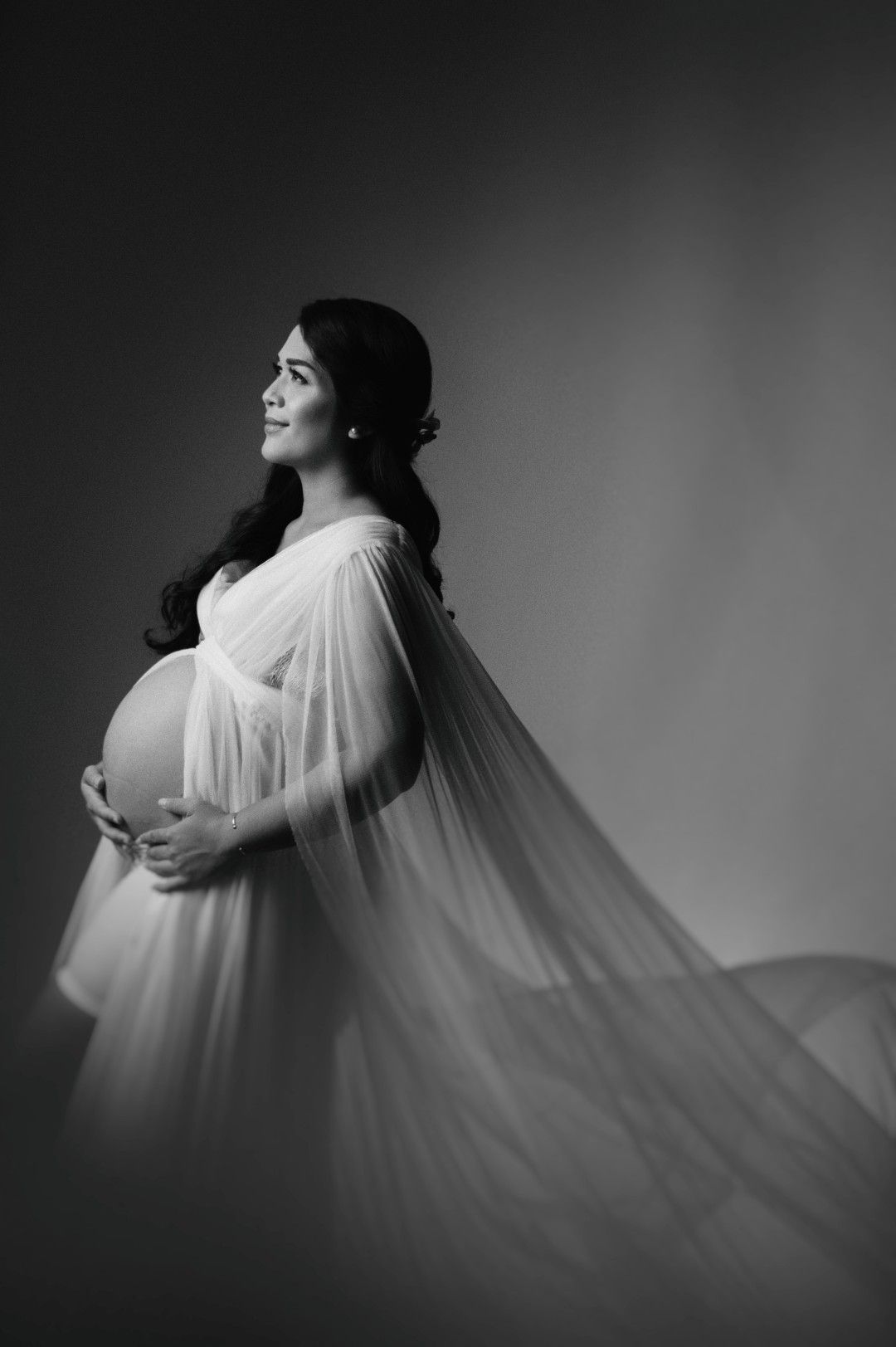Black and white maternity photo of a smiling pregnant woman in a flowing white sheer gown, cradling her baby bump.