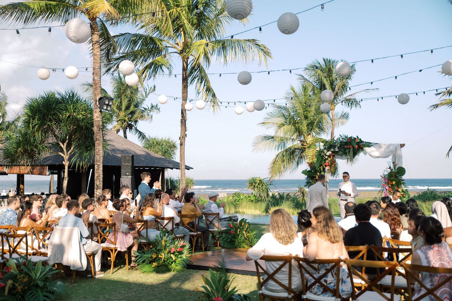 Outdoor beach wedding ceremony with guests seated under string lights and palm trees by the ocean.