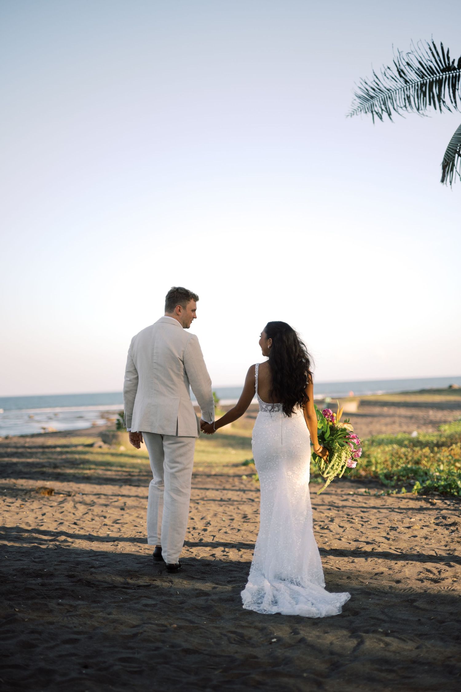 Bride and groom hold hands walking toward ocean at sunset on black sand beach.