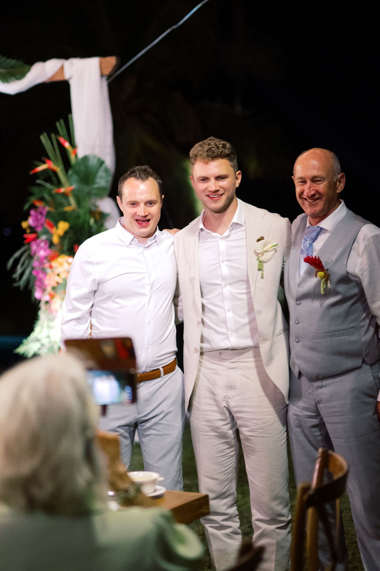 Groom poses with two men in white shirts and grey suits at evening wedding.