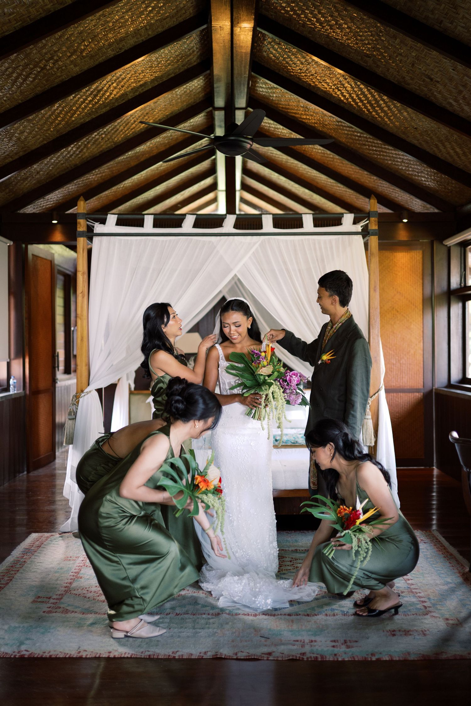 Bride in white gown surrounded by bridesmaids in green dresses inside a Balinese villa.