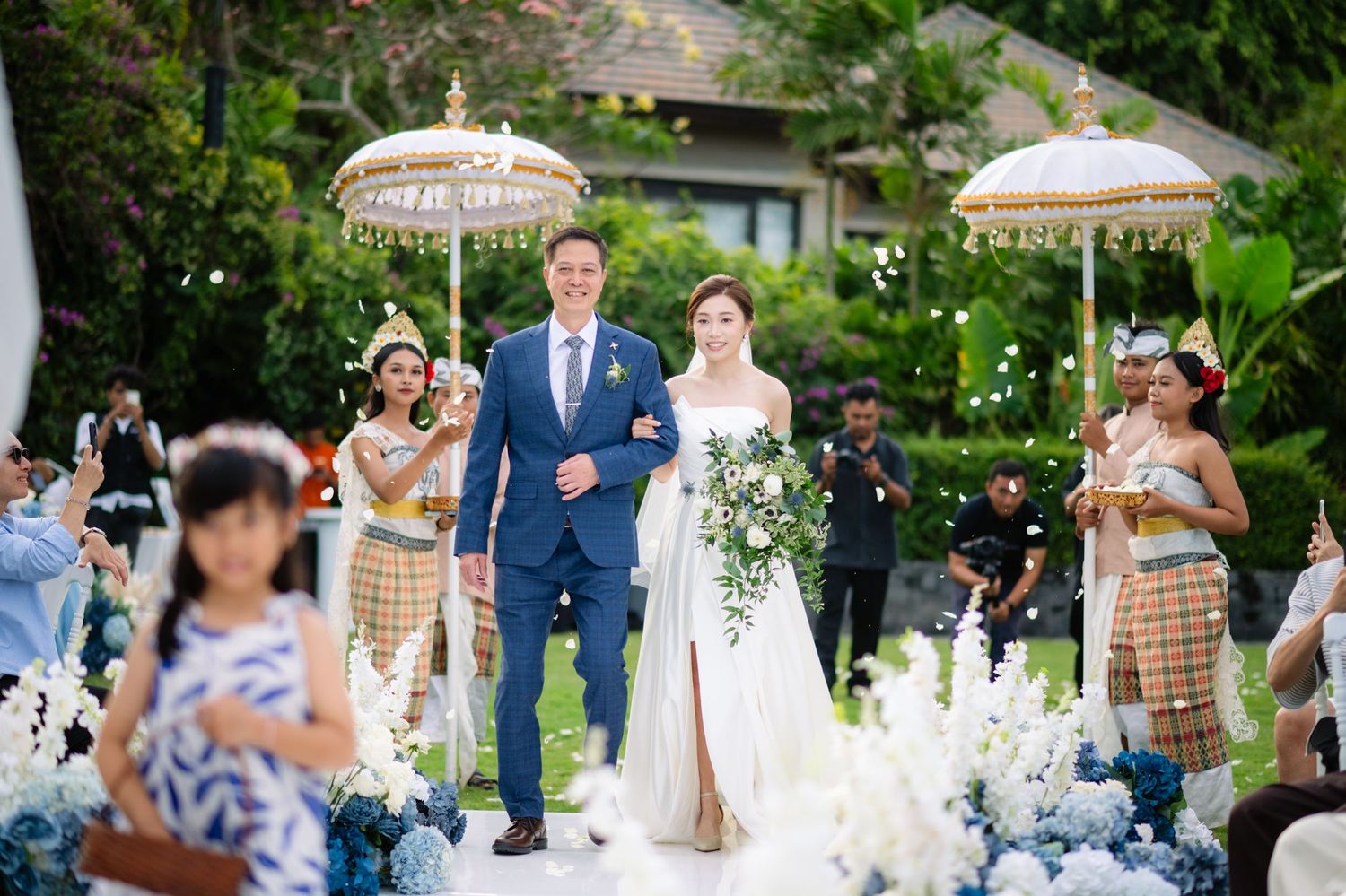 Bride in a white gown and groom in a navy suit walk down a flower-lined aisle at a tropical outdoor wedding as guests and attendants watch.