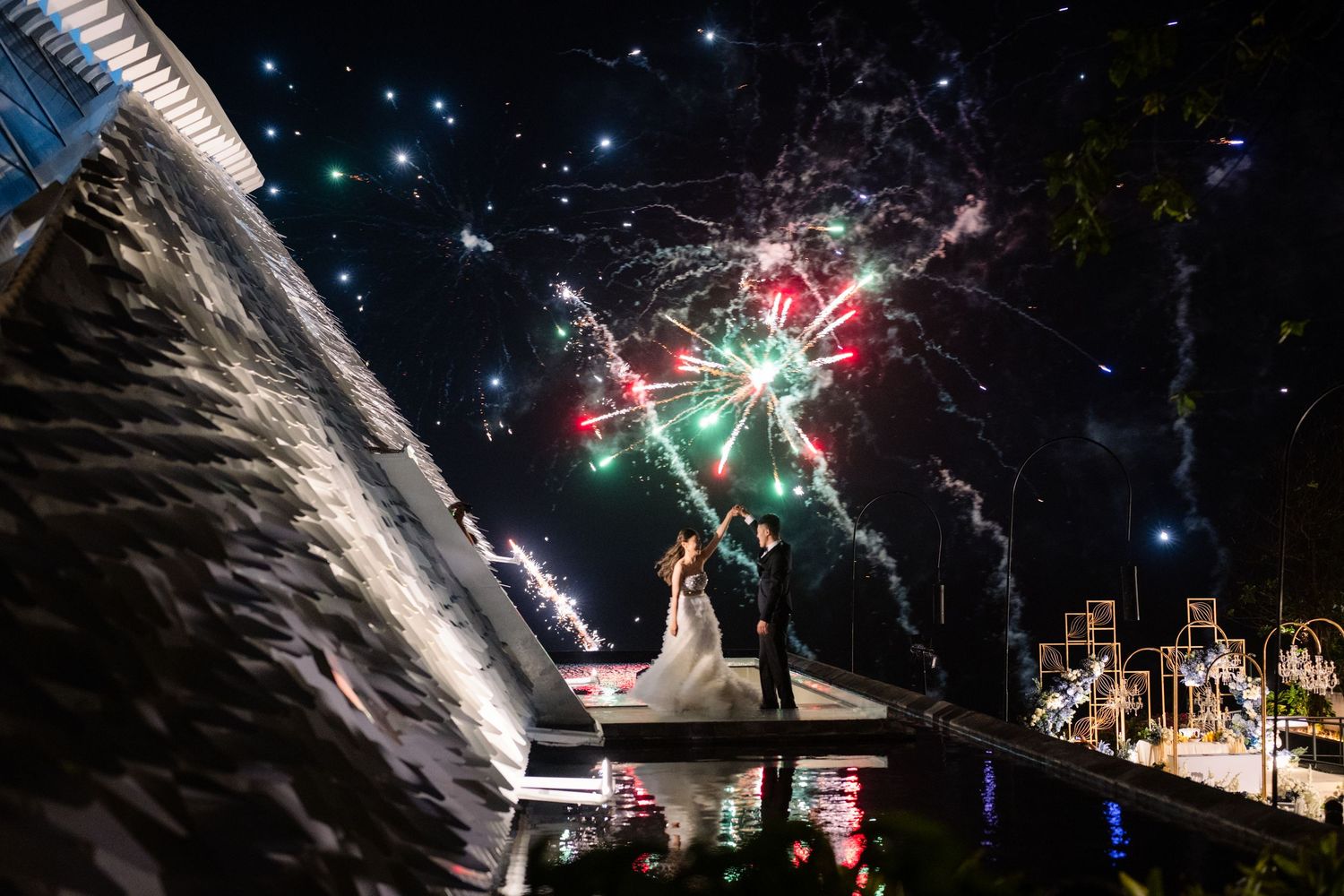 Bride and groom embrace on a waterfront platform at night as colorful fireworks burst across the dark sky above them.