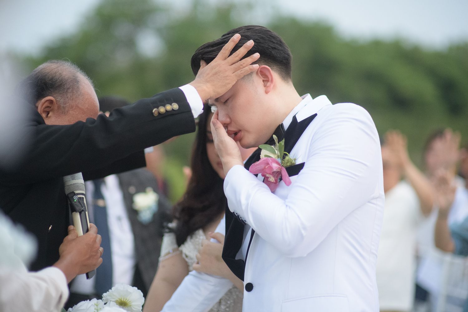 Emotional groom in white suit crying during outdoor wedding ceremony while officiant places hand on his head.