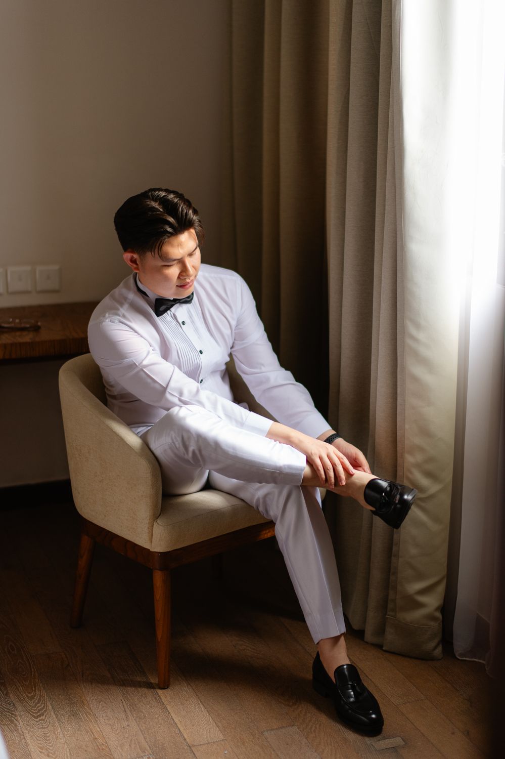 Young man in white tuxedo shirt and gray trousers adjusting his shoe while seated in a beige chair.