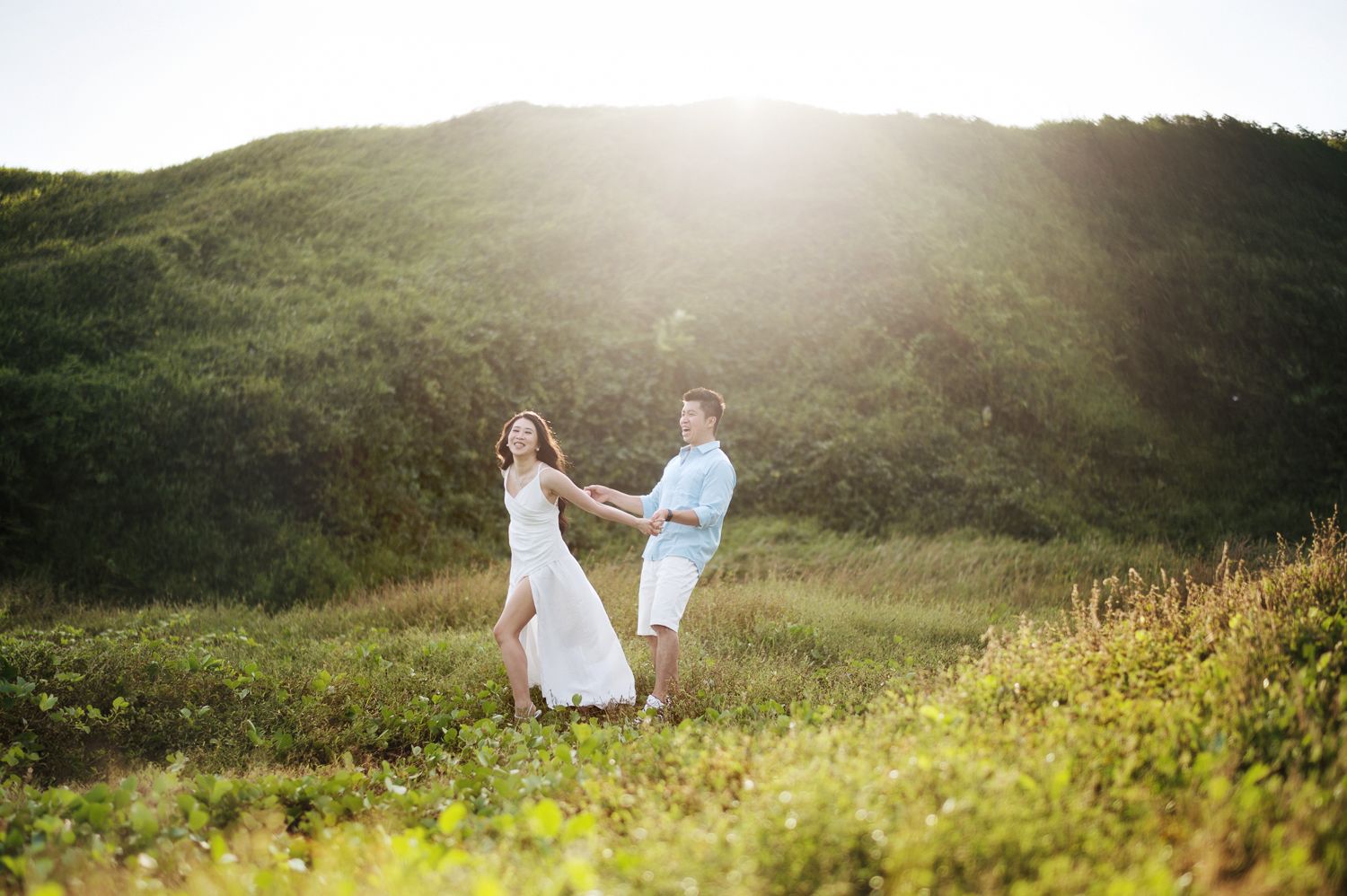 Couple holding hands and dancing in sunlit green field with glowing hillside backdrop.