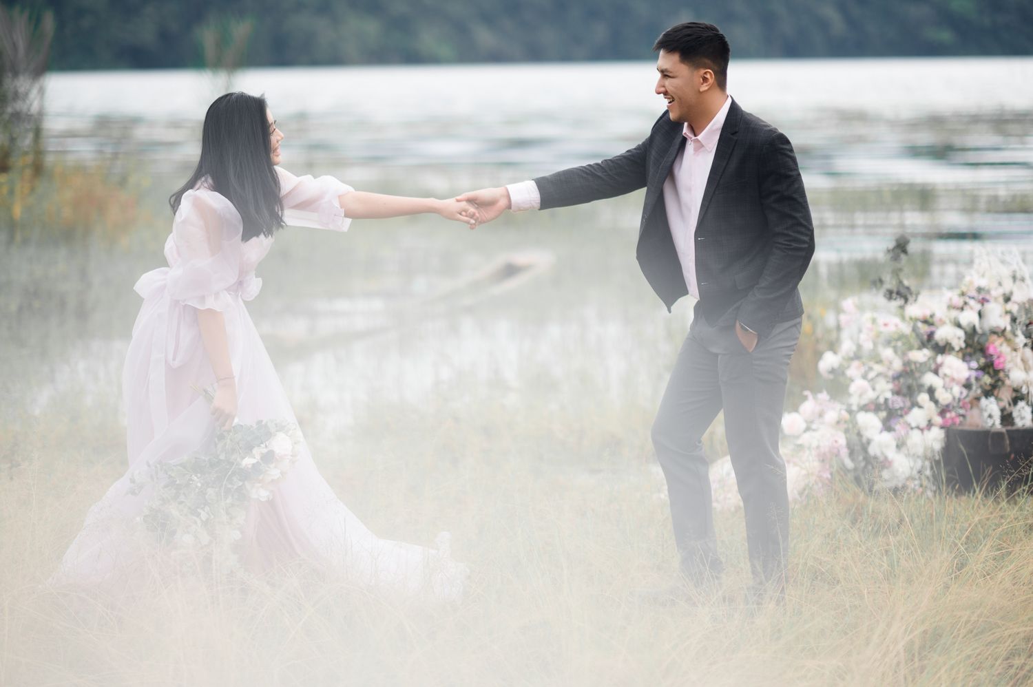 Couple holding hands in a romantic outdoor wedding photoshoot near a lake, surrounded by tall grass and floral arrangements.