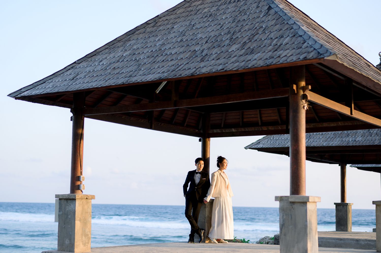 Couple in wedding attire standing under an elegant open-air pavilion overlooking the ocean at a beachside ceremony.