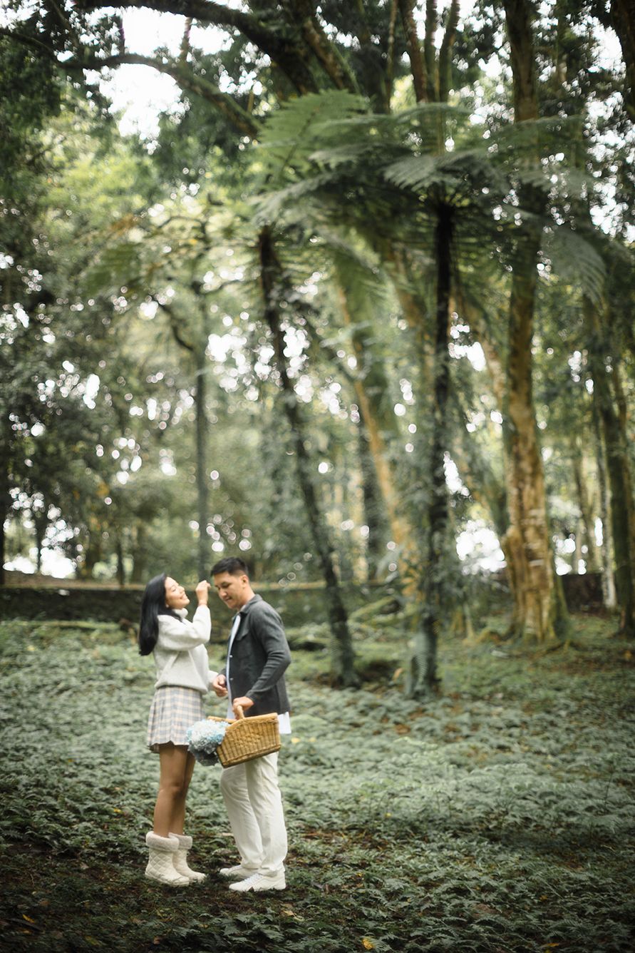 Couple sharing a romantic moment in a lush green forest, man holding a wicker basket while woman touches his face.