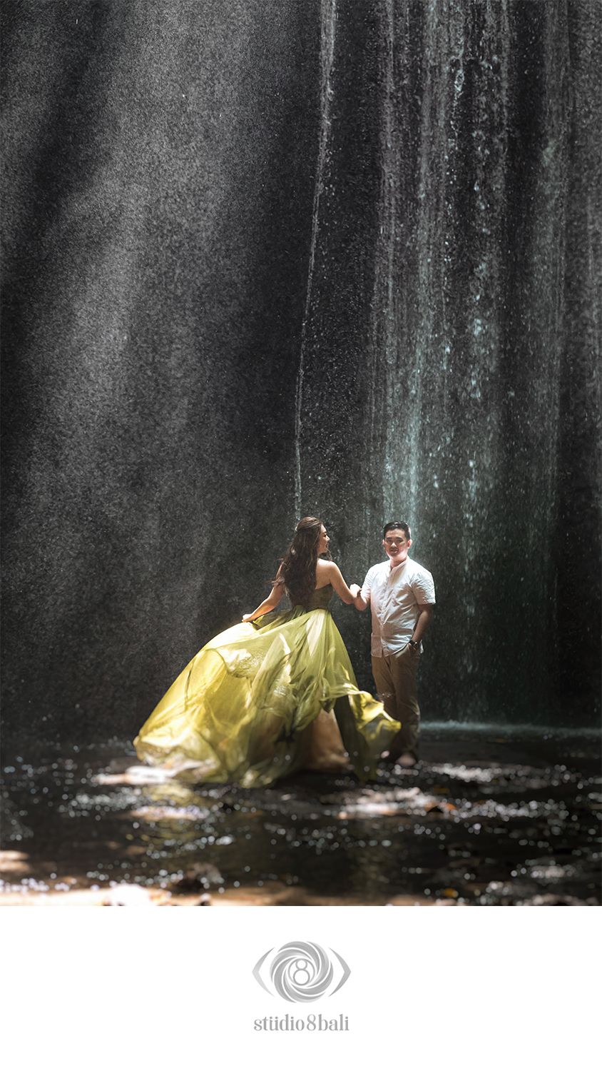 Couple in romantic pose near waterfall, woman in flowing yellow gown, man in white shirt, Studio 8 Bali photography.