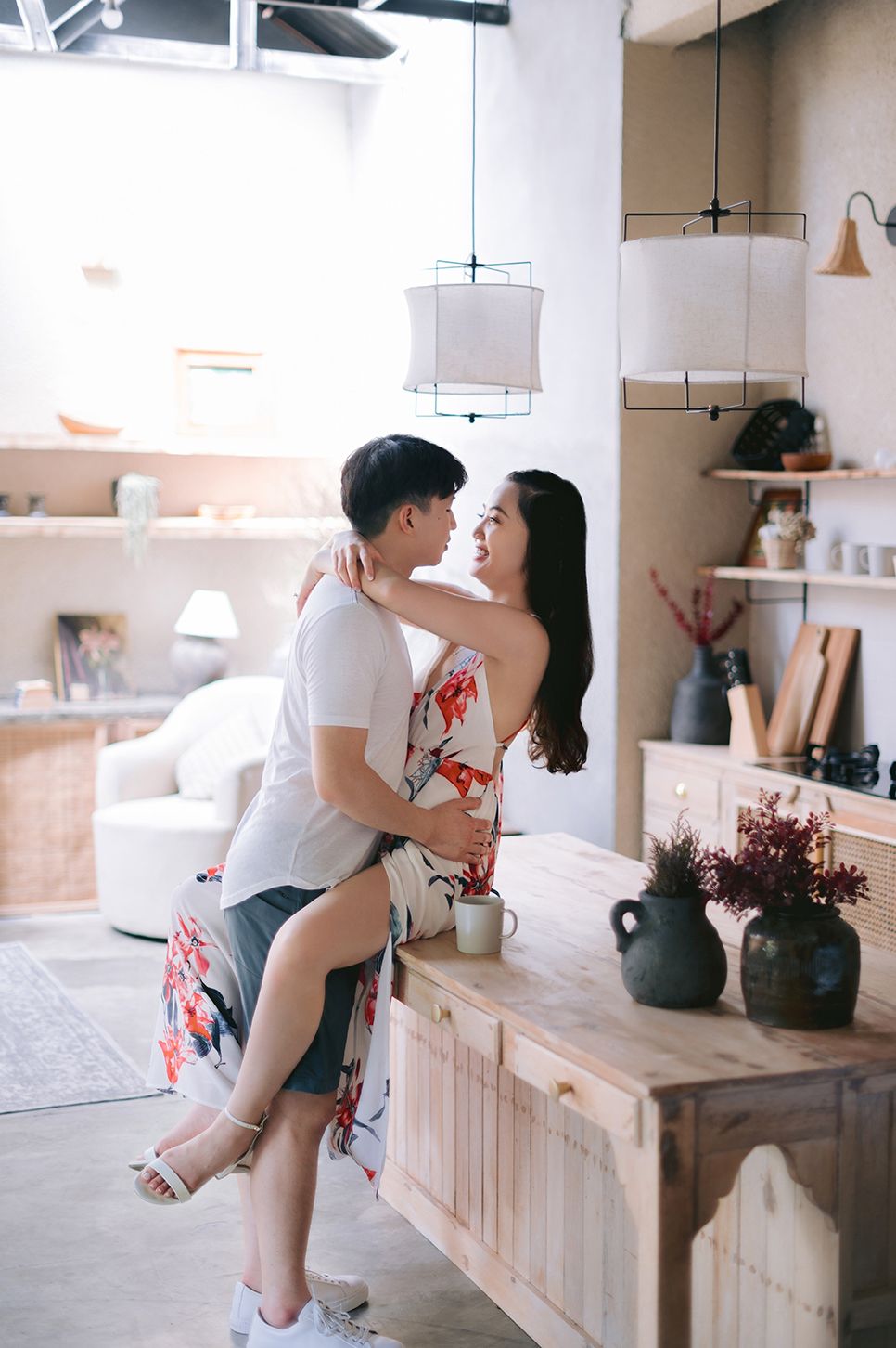 A couple shares a romantic embrace in a cozy, warmly decorated kitchen, with the woman seated on a wooden counter.