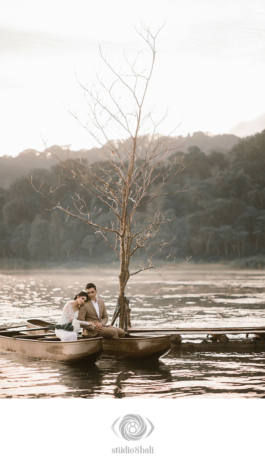 Couple sitting in a wooden boat on a serene lake with a bare tree, misty mountains in the background.