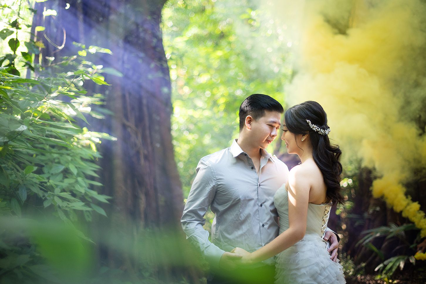 Couple in wedding attire sharing romantic moment in lush forest with yellow smoke bomb backdrop.