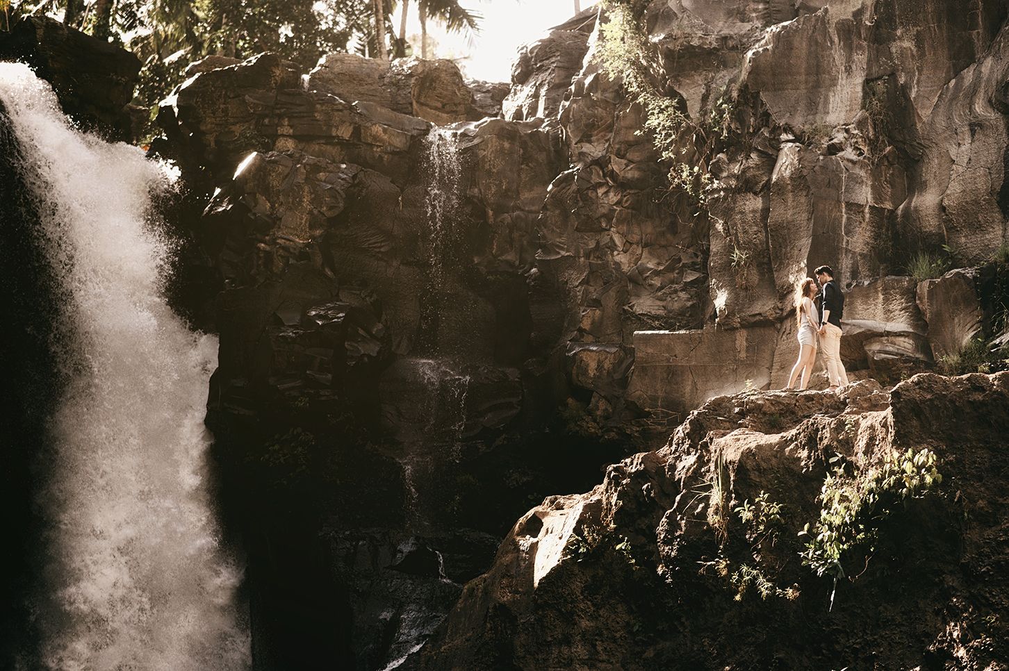Couple standing on rocky cliff beside a dramatic waterfall surrounded by lush tropical greenery.