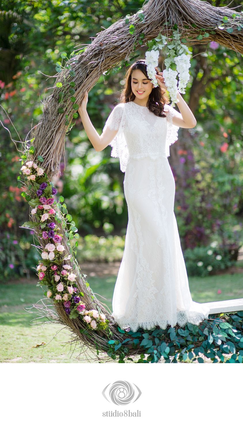 Bride in white lace gown with cape sleeves poses under floral twig arch with pink and purple blooms in garden.