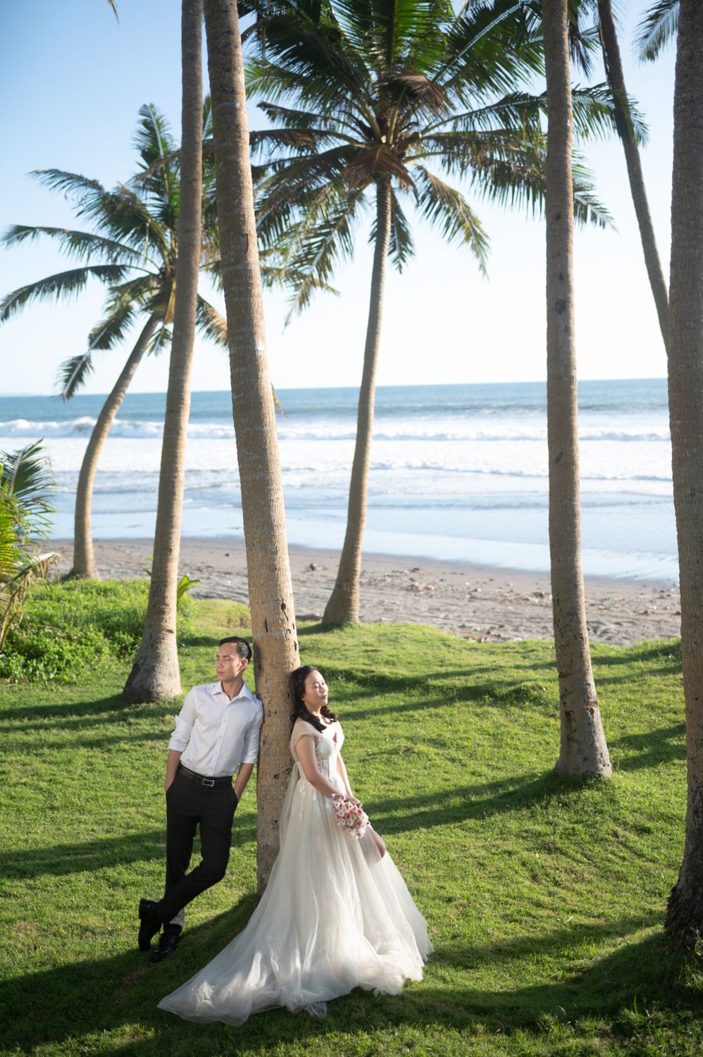 Couple in wedding attire posing among palm trees on a tropical beach with ocean waves in the background.