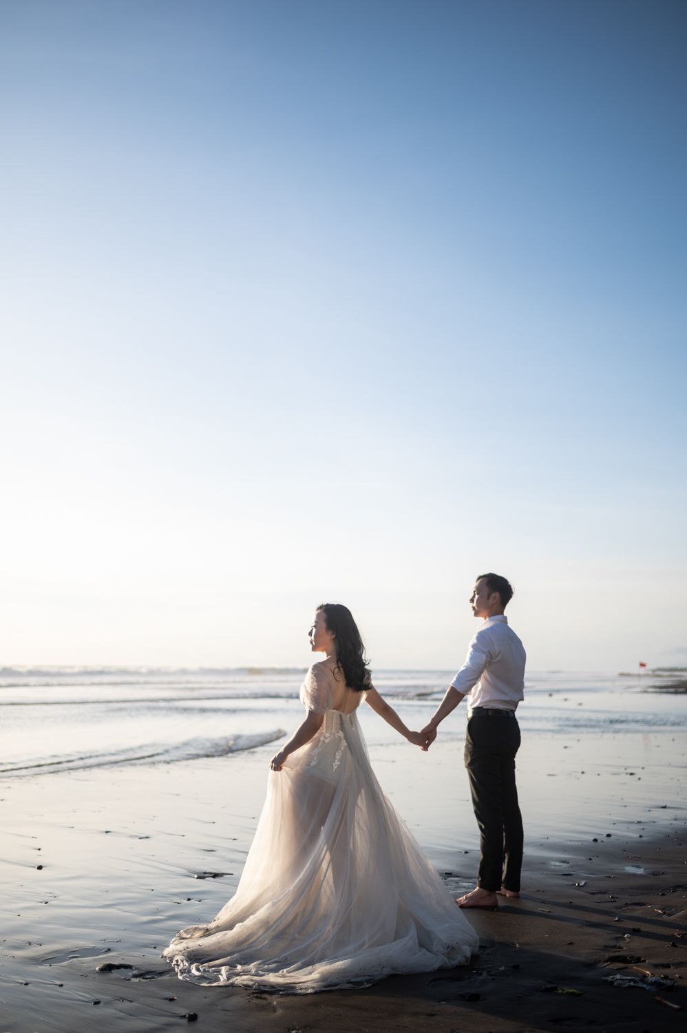 Romantic beach wedding couple holding hands at sunset with ocean waves and clear blue sky.