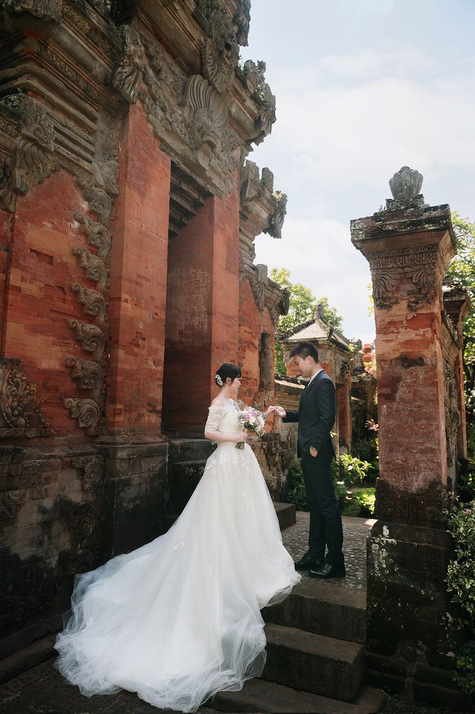 Bride and groom exchange vows at ornate Balinese temple, bride in flowing white gown with long train on stone steps.