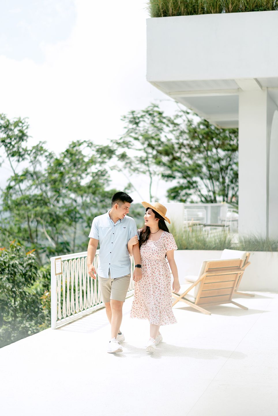A couple walks hand-in-hand on a bright terrace, smiling at each other, woman in floral dress and hat, man in casual attire.