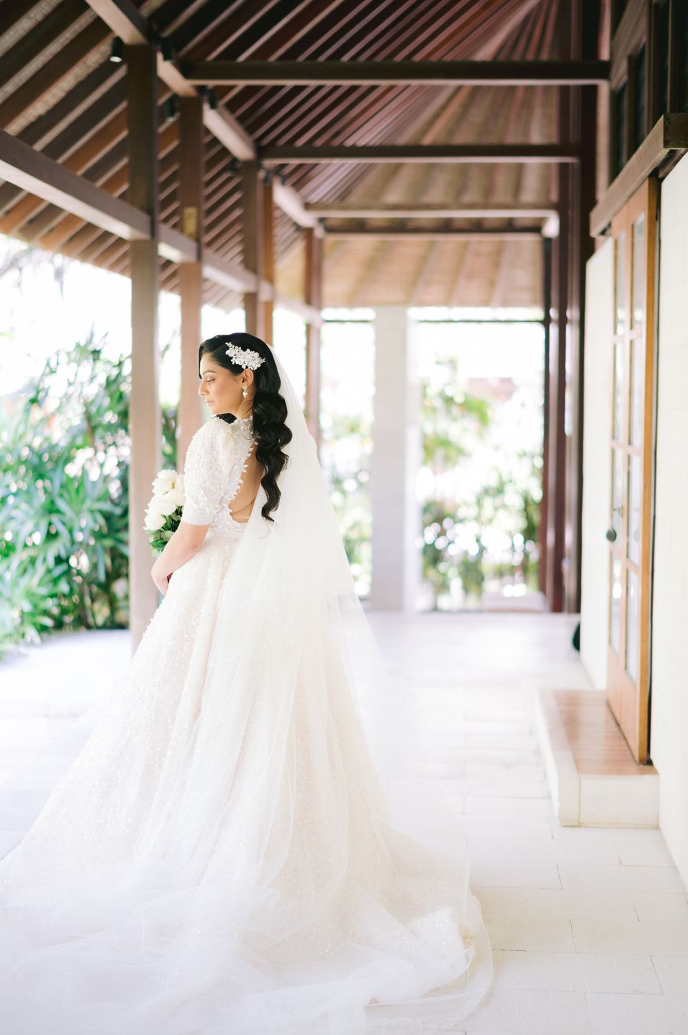 Bride in elegant white ball gown with lace sleeves holding bouquet in tropical venue corridor.