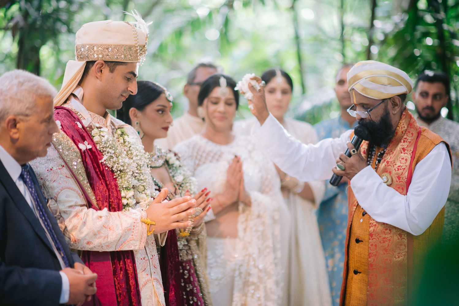 Traditional Indian wedding ceremony with groom in red sherwani and bride in white, officiant blessing couple outdoors.