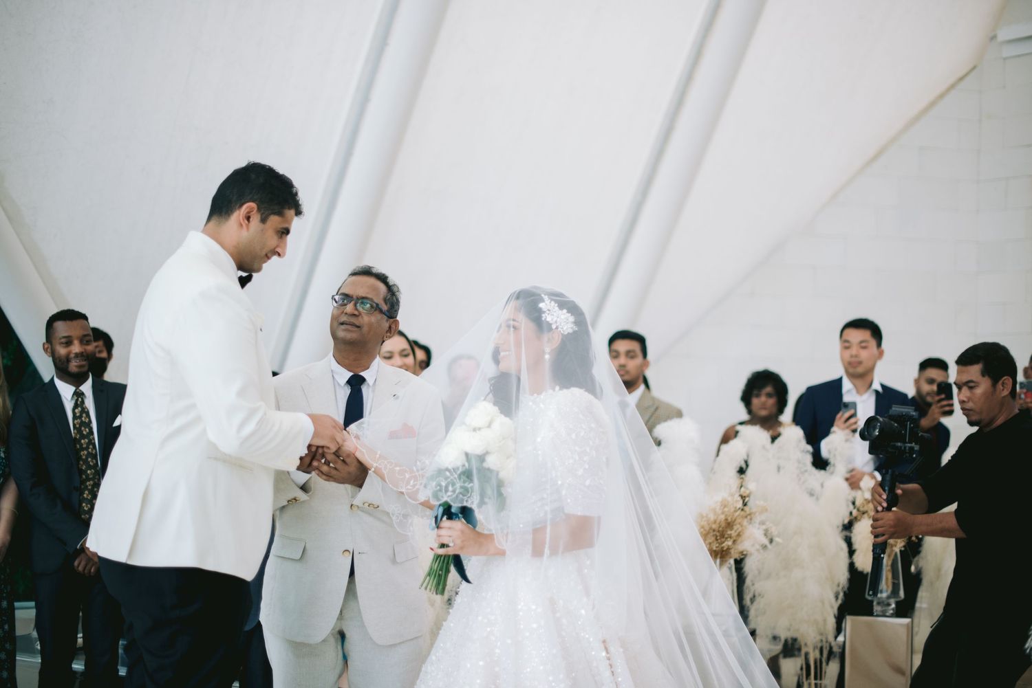 A bride in a white gown and groom in a white suit exchange vows at an elegant wedding ceremony under a tent.