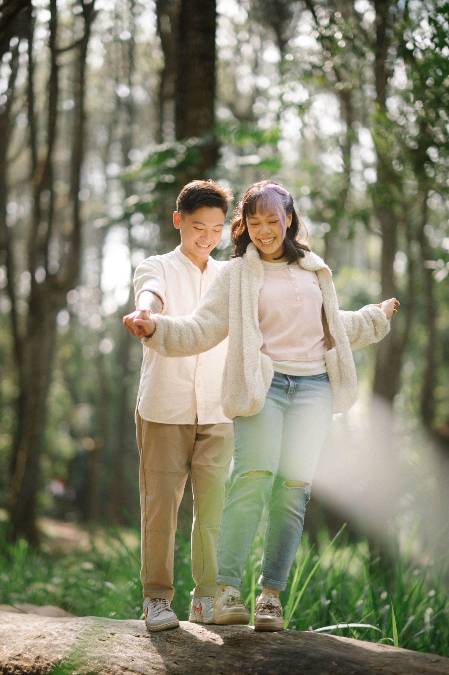 Young Asian couple smiling and holding hands while balancing on a log in a sunlit forest park.