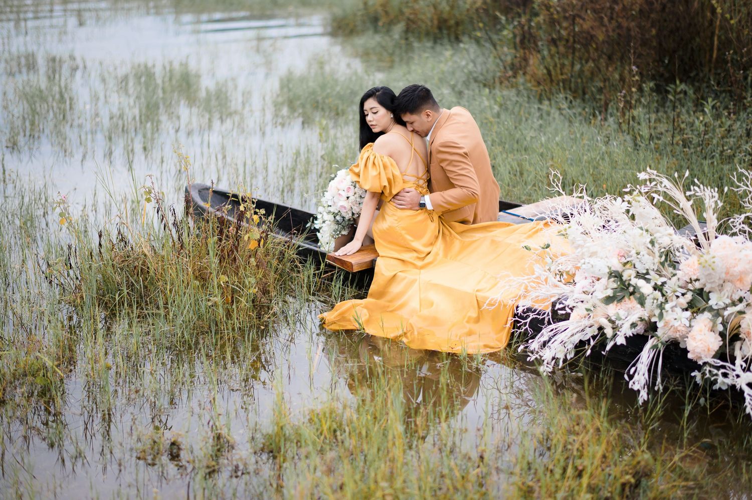 Couple in yellow and brown outfits sitting romantically in a flower-adorned boat on a serene marshy lake.